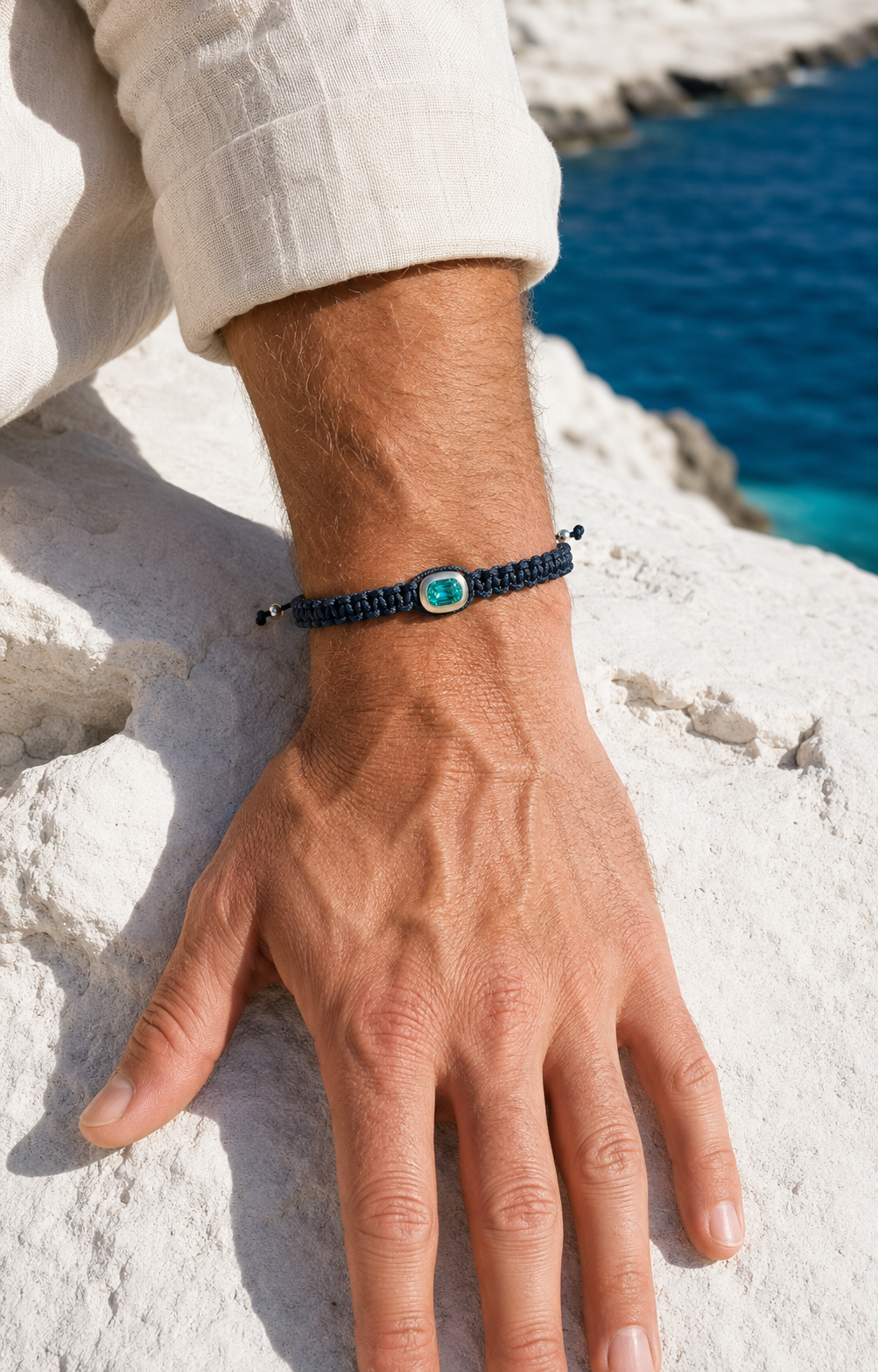 The REBL Signature Bracelet on a sunlit forearm against white Mediterranean stone, the blue braided cord and light-blue Paraíba stone catching the light.
